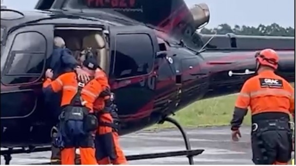 Foto de uma pessoa resgatada entrando num helicóptero, amparada por dois profissionais. 